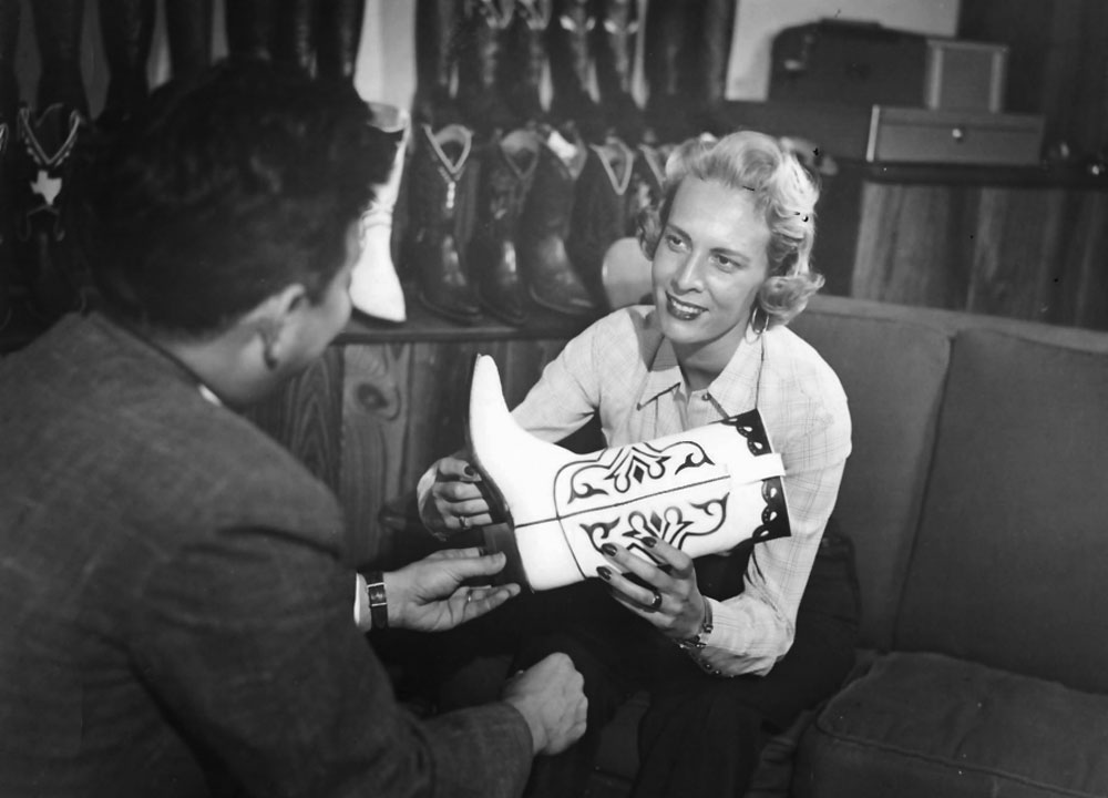 Una fotografía en blanco y negro de una joven Jane Chilton Justin con cabello rubio, vestida con traje de negocios y sosteniendo una bota de vaquero, presentándola a un cliente.
